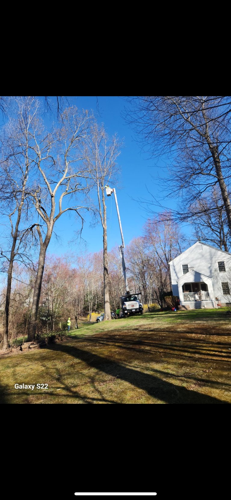 Utility work truck with tall lift equipment next to a bare birch tree in front of a white colonial house on a sunny day