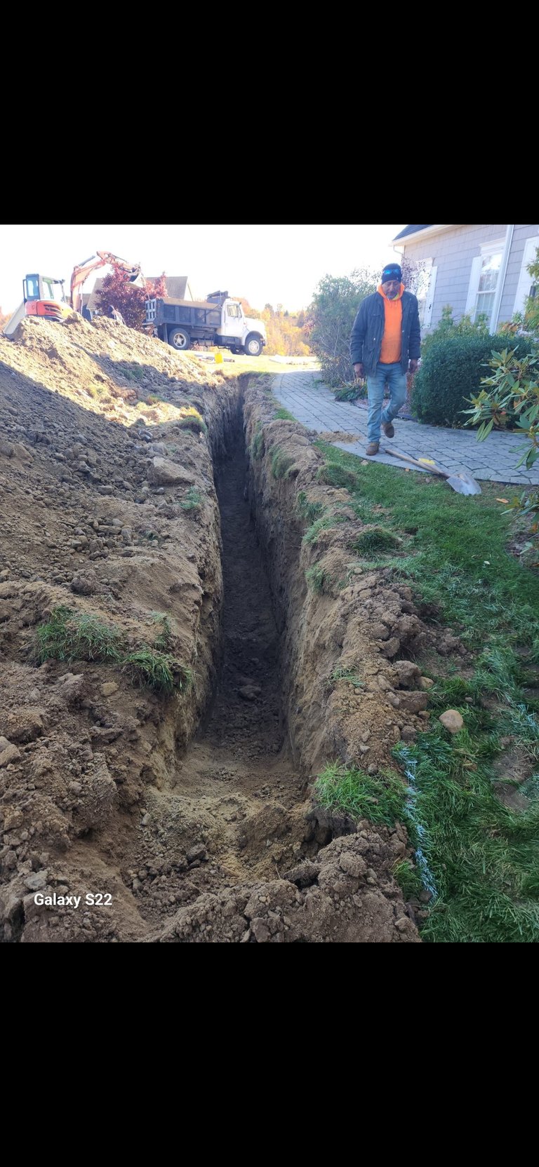 Construction site showing deep trench excavation with worker in safety gear standing nearby and heavy machinery in background