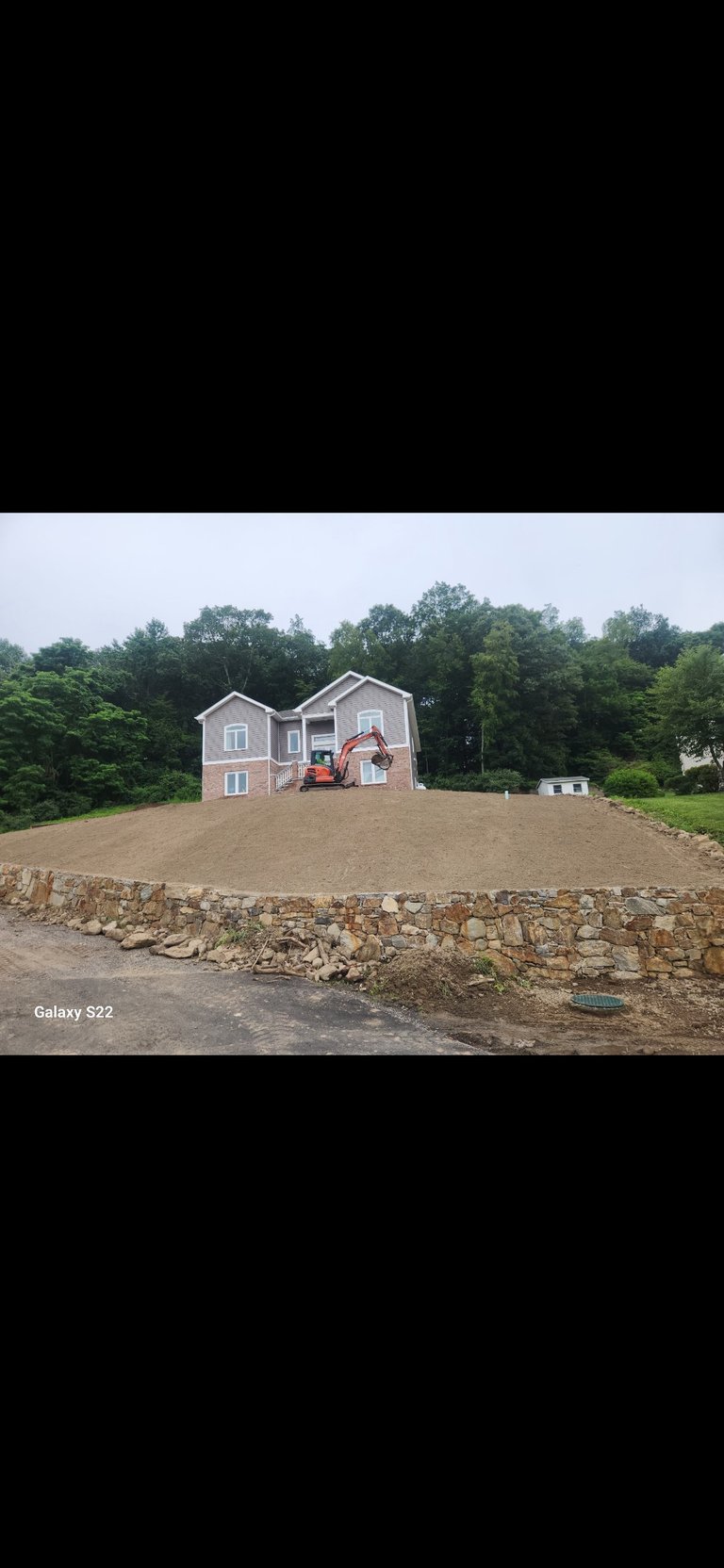 Two-story residential house under construction with orange excavator on unpaved driveway, surrounded by trees
